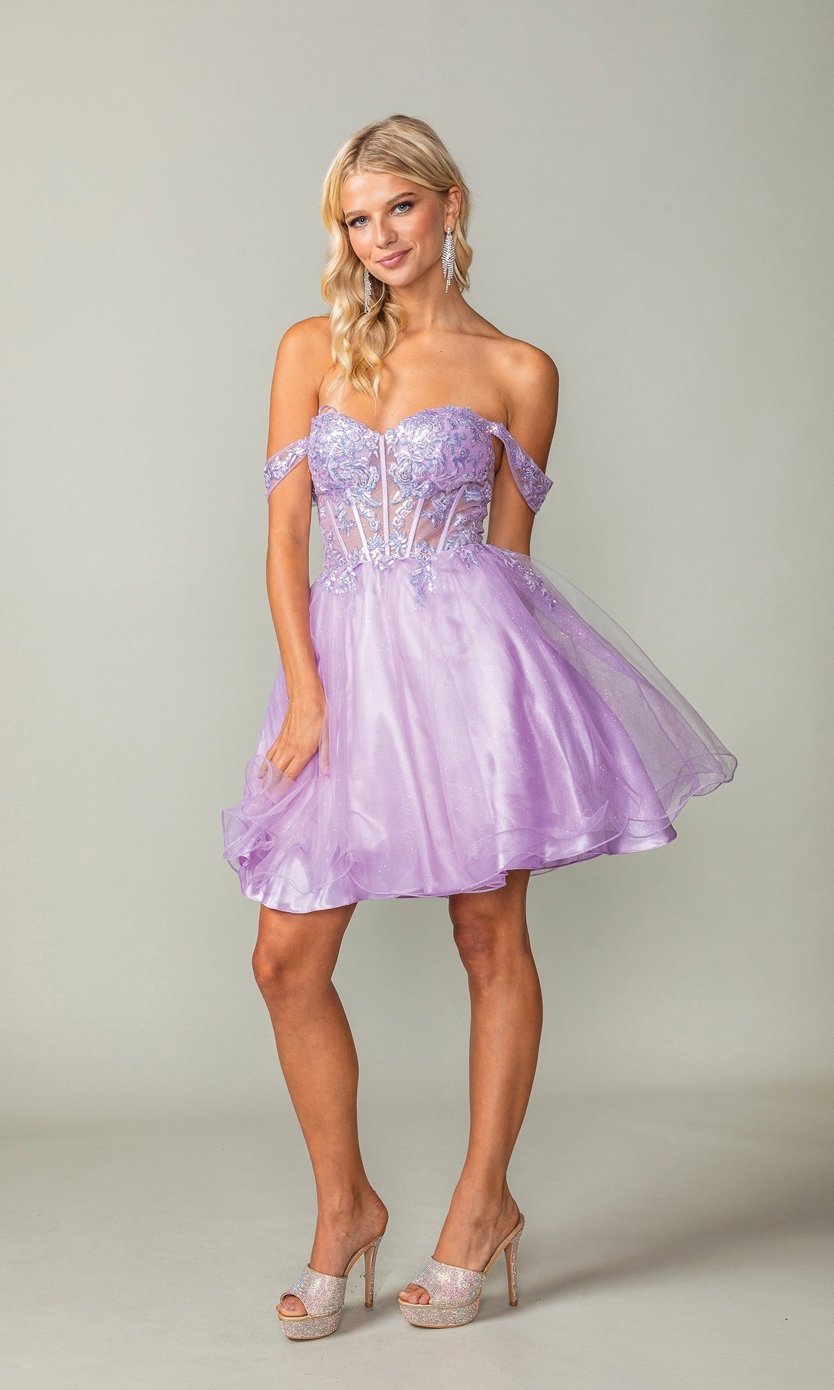 A woman models the Dancing Queen 3383 Short Corset Homecoming Dress, featuring an off-the-shoulder lavender sequin bodice and flared tulle skirt, paired with high heels against a plain backdrop.